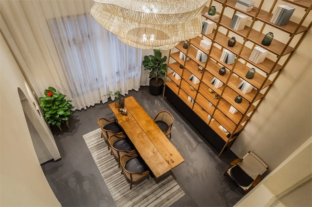 A minimalist living room with a wooden table, chairs, and bookshelf, featuring light-colored textiles and plants.