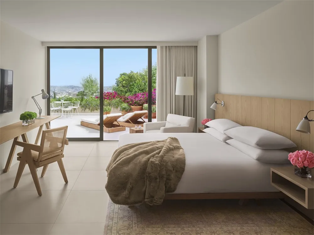 Modern bedroom with white walls, glass door to balcony, wooden bed frame, white bedding, and beige blanket.