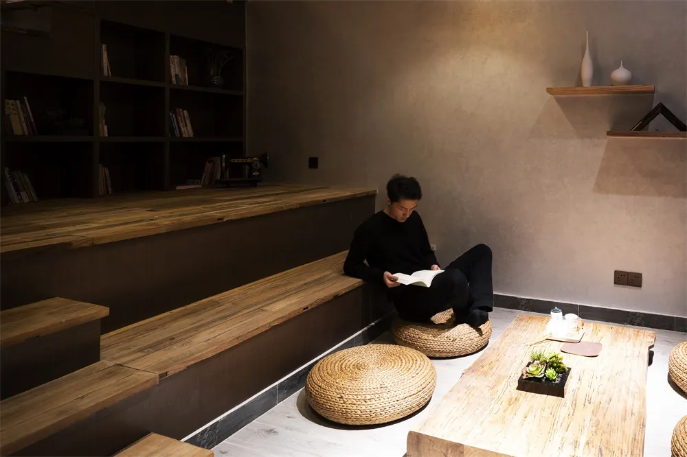 一位人士坐在編織坐墊上,置身於燈光昏黃、木質元素充滿的室內空間,正在閱讀一本書。