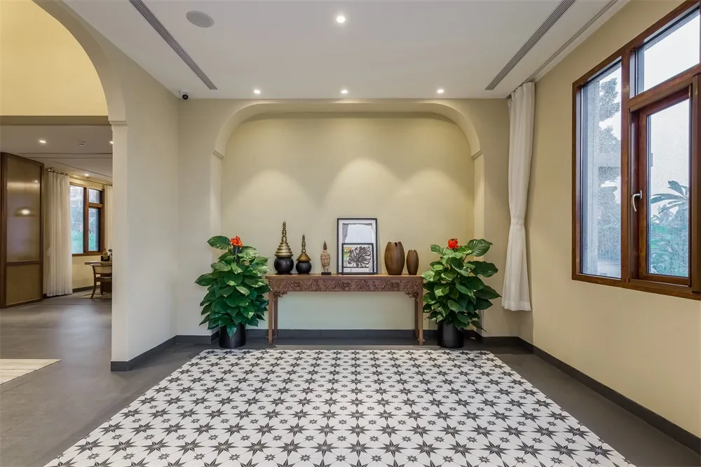 A modern living room featuring a patterned carpet, a wooden console table with decorative items, green plants, and large wind