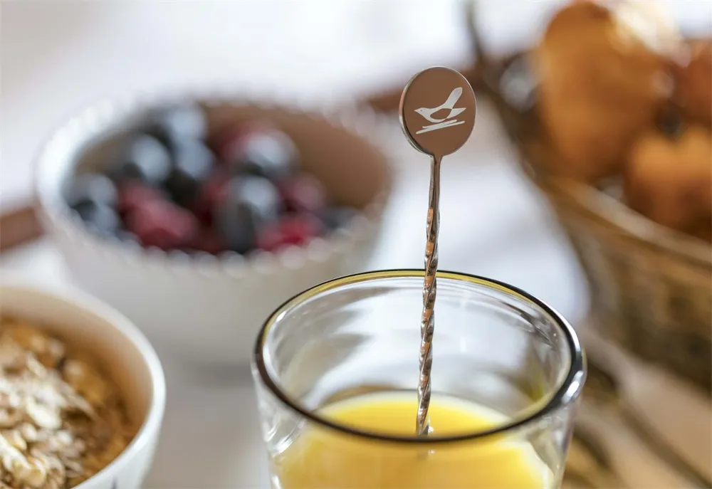 Close-up of a minimalist breakfast scene with a metal stirring spoon in a glass of orange juice, a bowl of mixed berries, and