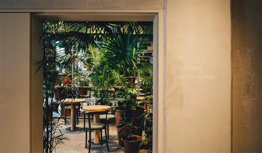 Industrial restaurant interior with concrete walls, large plants, and rustic furniture.