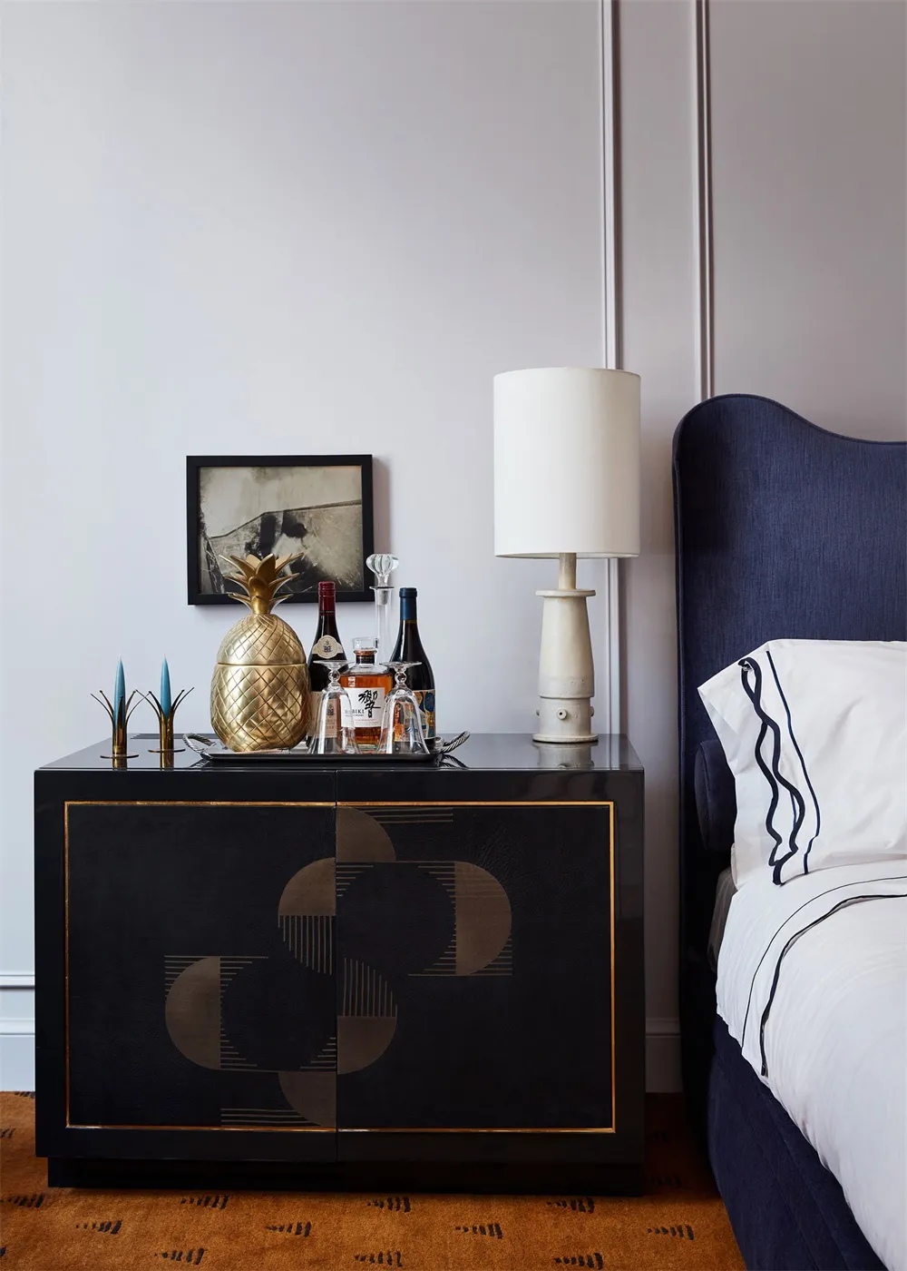 A modern bedroom corner with a black sideboard featuring a brass pineapple ice bucket, a framed artwork, a table lamp, and a