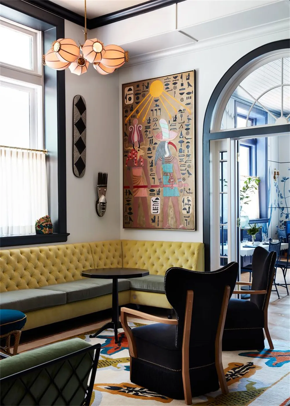 A modern restaurant lounge with a yellow upholstered sofa, black wingback chairs, and a colorful patterned carpet.