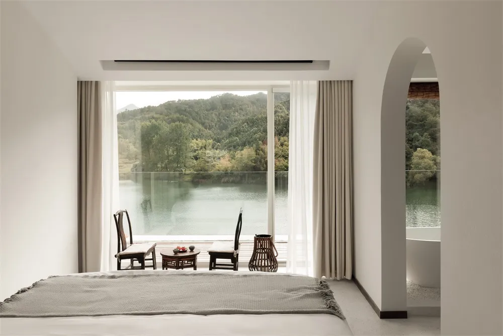 A modern, minimalist bedroom featuring white walls, wood furniture, and a glass window overlooking a scenic lake and mountain