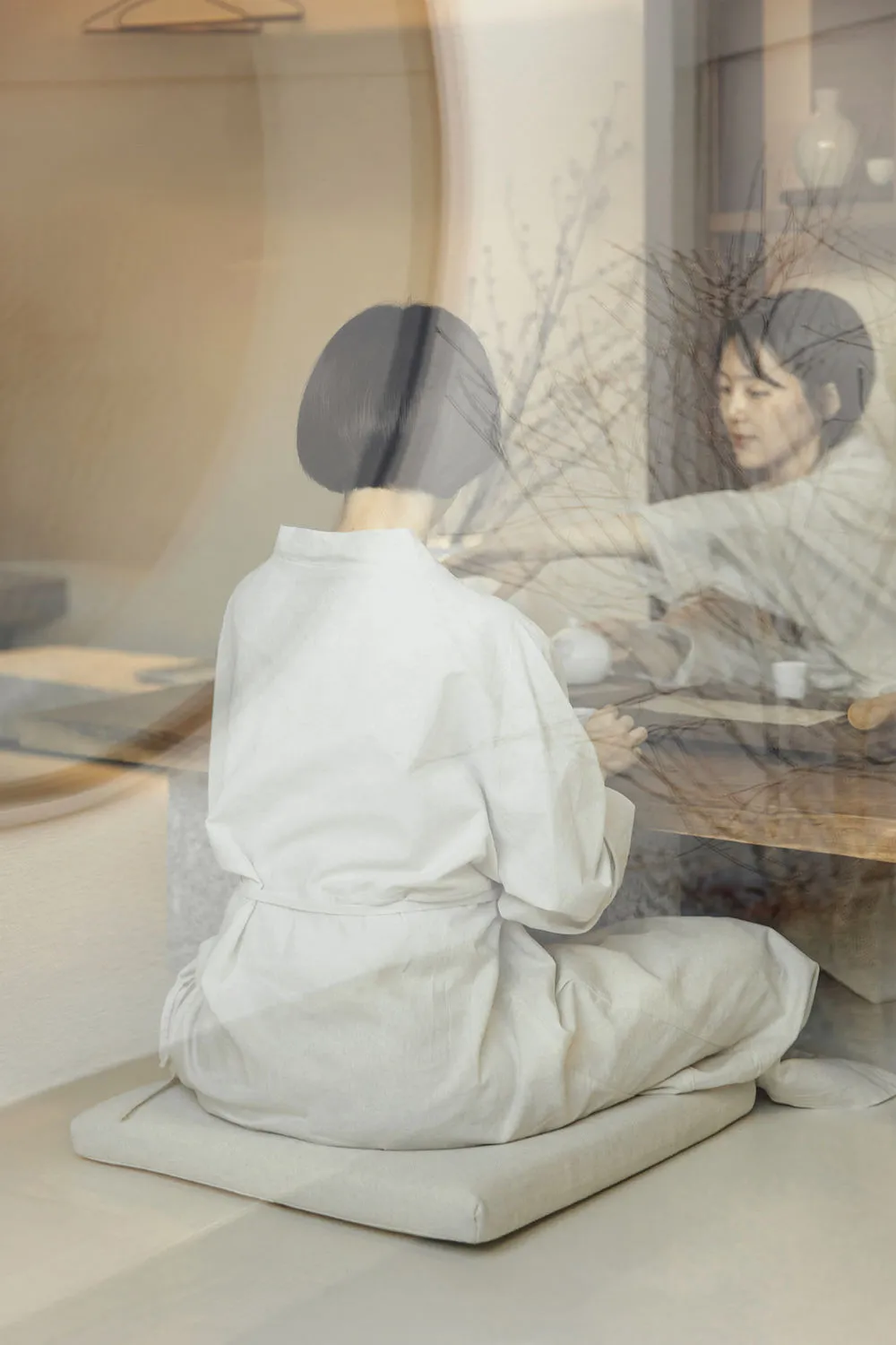 A person dressed in white textiles performs a tea ceremony in a serene, minimalist setting.