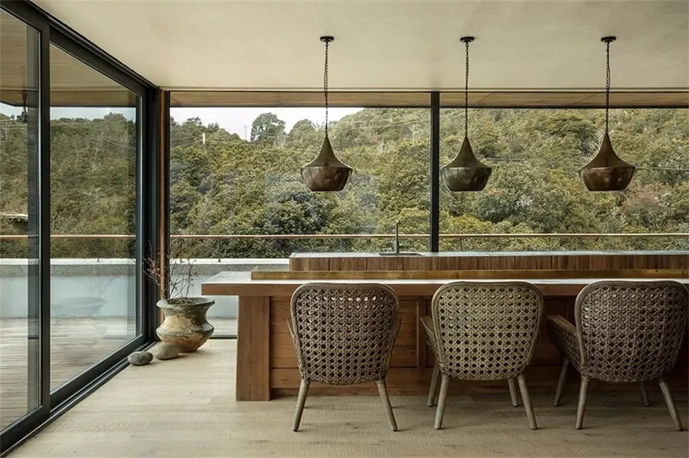 A modern kitchen with a wooden counter, wicker chairs, and large windows overlooking a forested area.