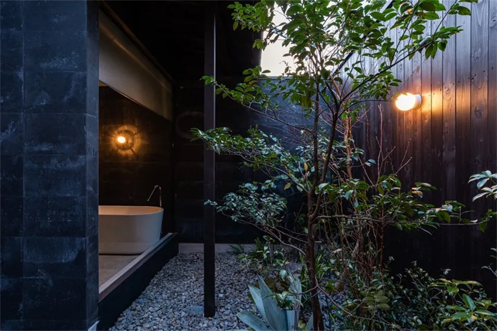 A minimalist bathroom with a concrete wall and a freestanding bathtub surrounded by greenery at dusk.