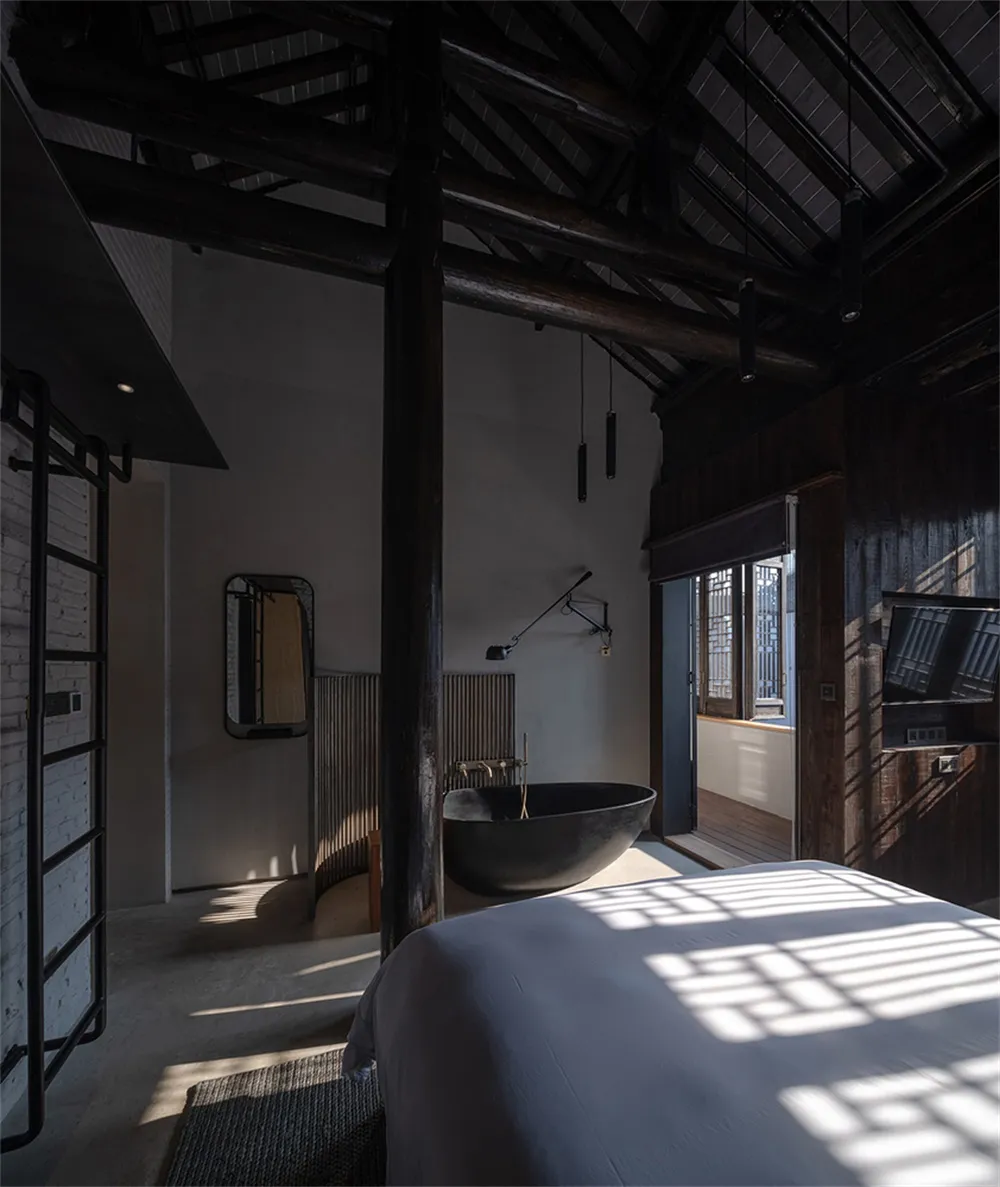 Interior of a minimalist bedroom with concrete walls and ceiling beams, featuring a black bathtub and a bed.