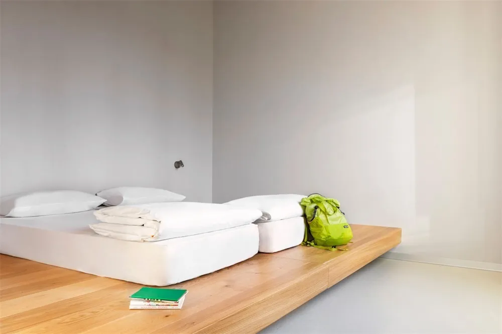 A modern minimalist bedroom with white bedding, a wooden floor, and a green backpack.