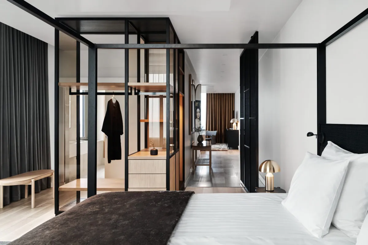 A minimalist bedroom with a bed, white bedding, and a glass-enclosed closet with wood shelves and metal accents.