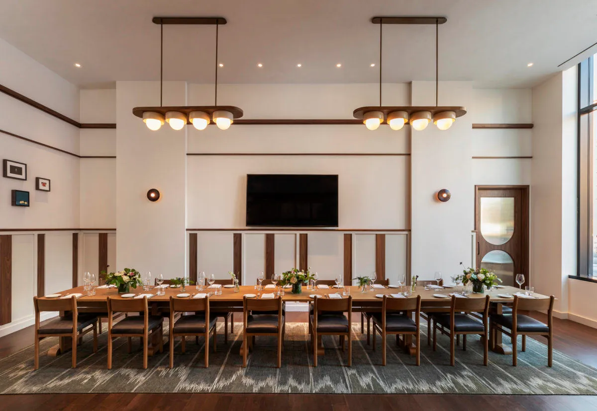 A modern dining room featuring a long, wooden dining table set for a large group, with pendant lights and a television in the