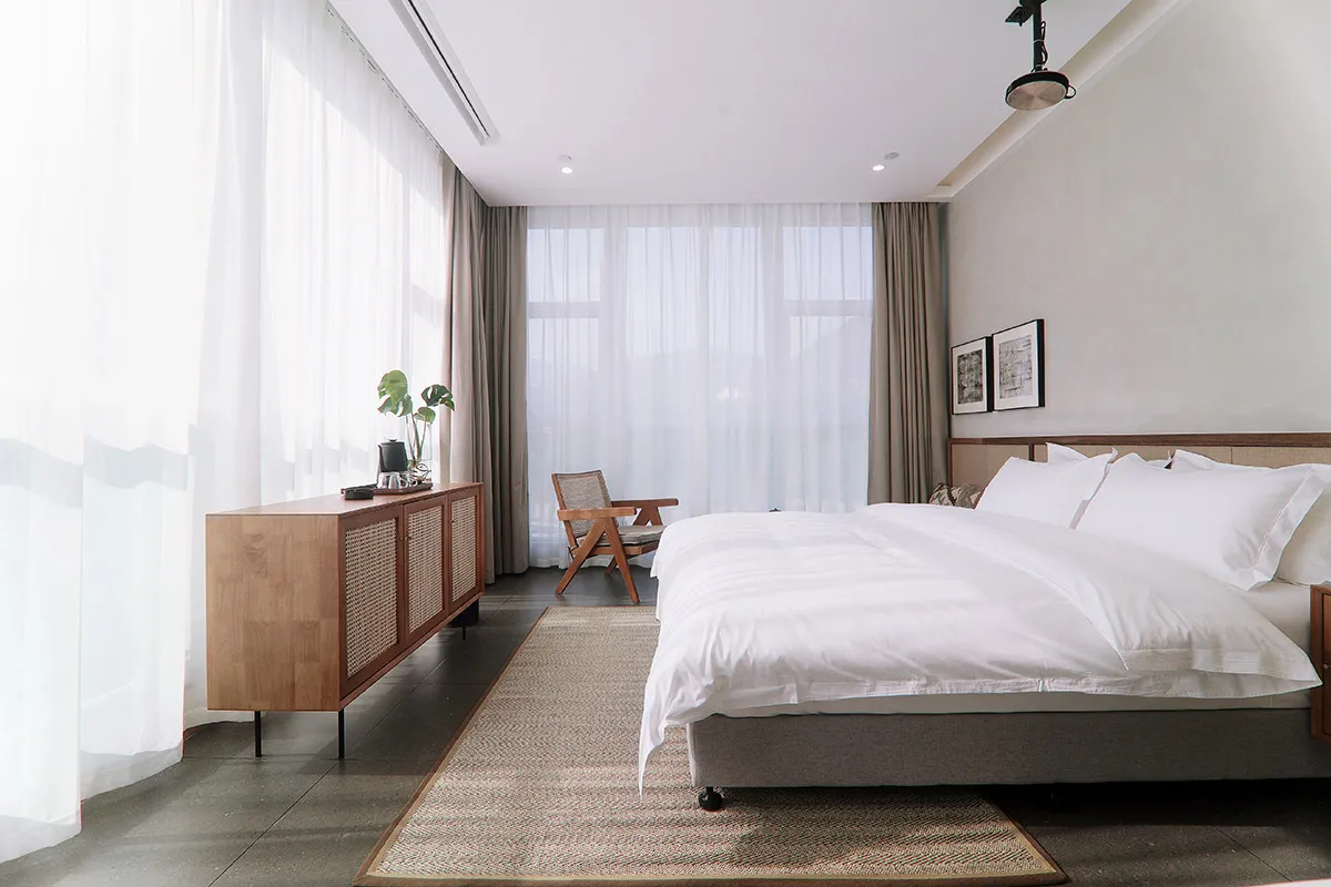 A modern, minimalist bedroom featuring a large window with white sheer curtains, a white bed with a beige headboard and white