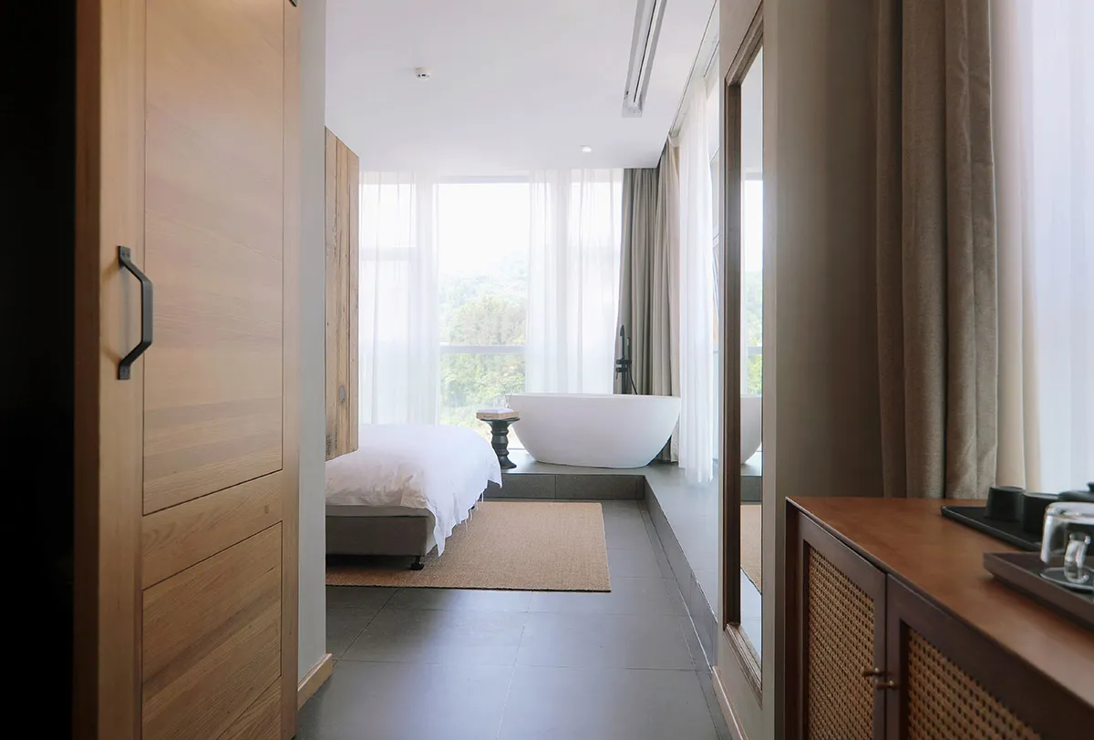 A minimalist bedroom with a wooden floor and concrete flooring, featuring a white bed and sheer curtains.