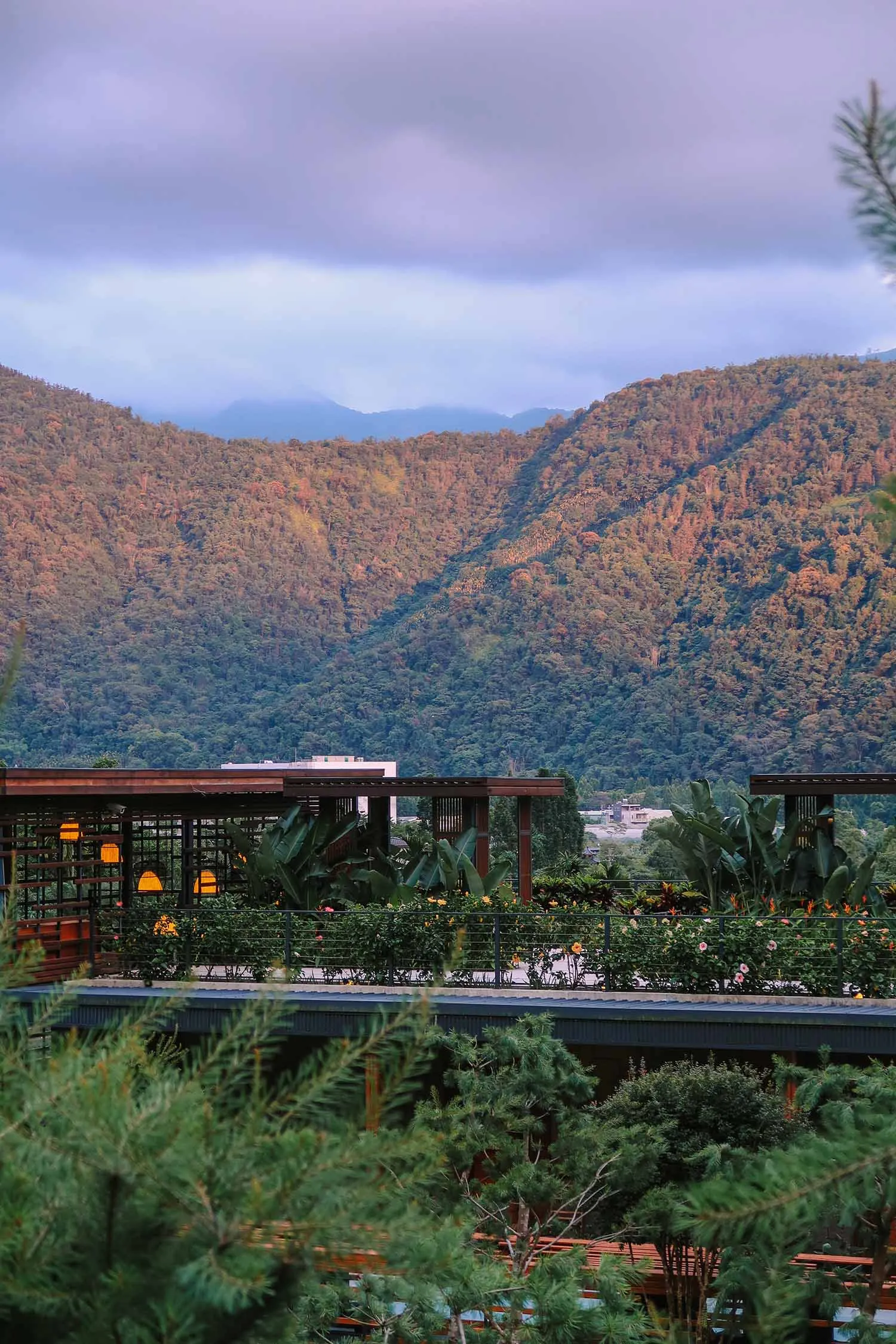 A scenic view of lush mountains with a cloudy sky in the background, framed by a wooden structure and green foliage.