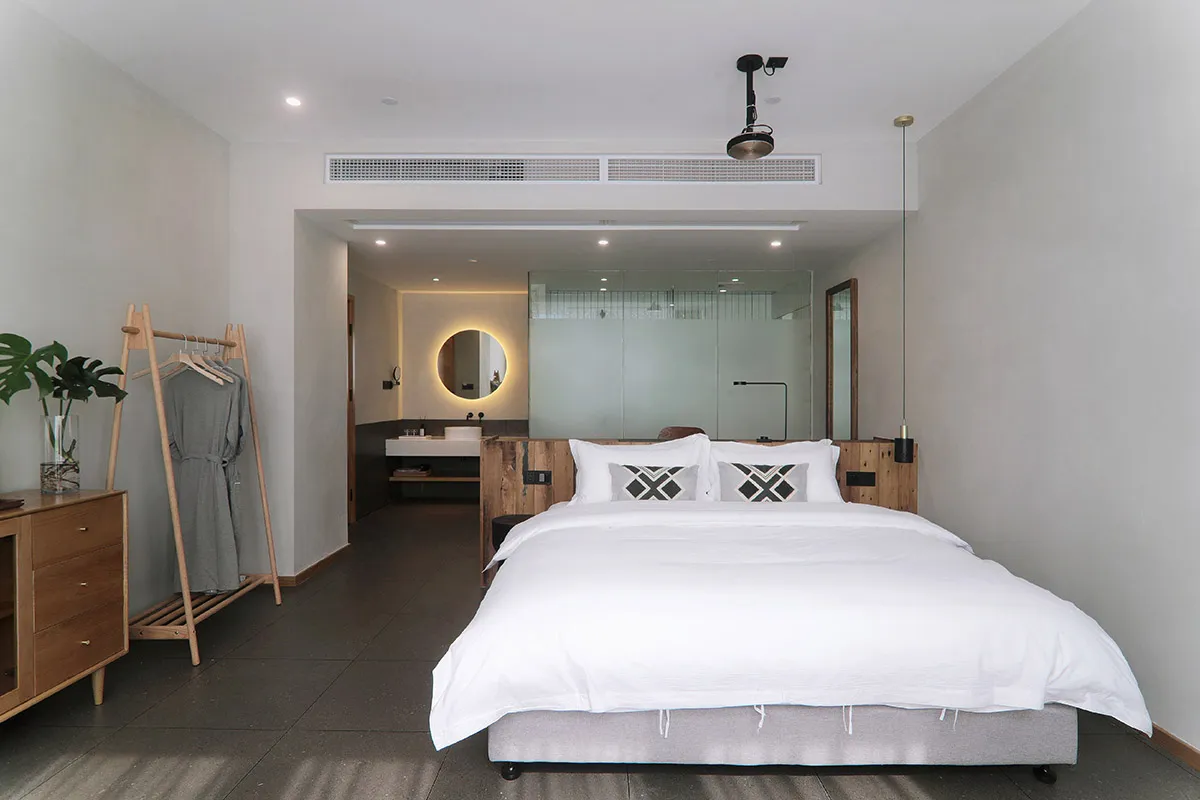 A minimalist bedroom featuring a white bed with geometric pillows, wooden headboard, and a glass partition in the background.
