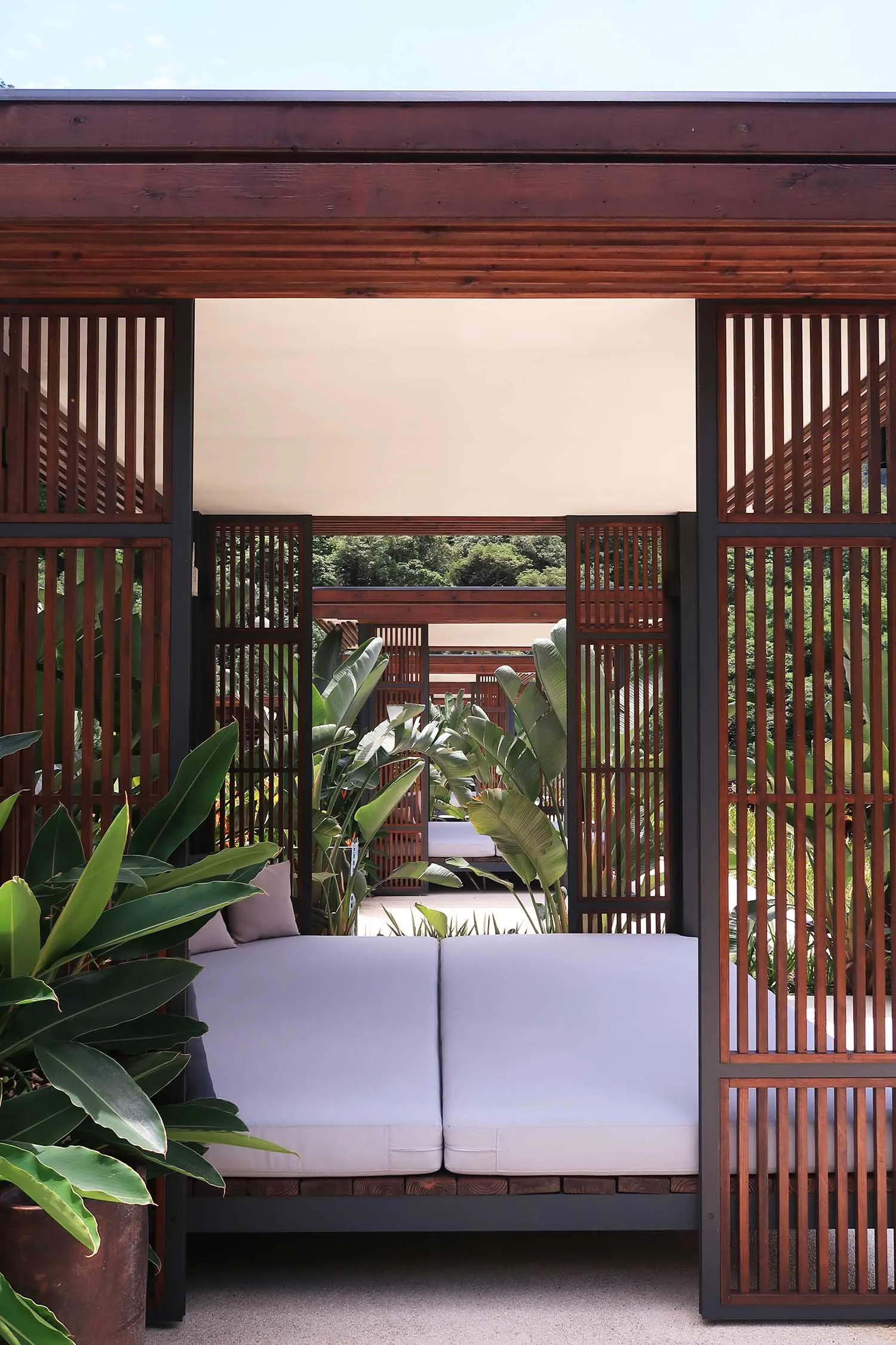 A modern outdoor lounge area with wooden framing and white cushions, surrounded by greenery.