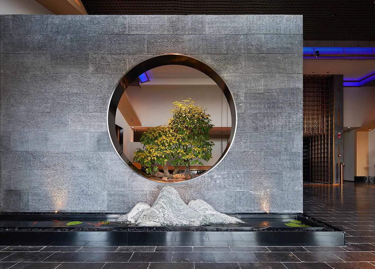 Modern hotel lobby with a circular window, tree, and water feature.
