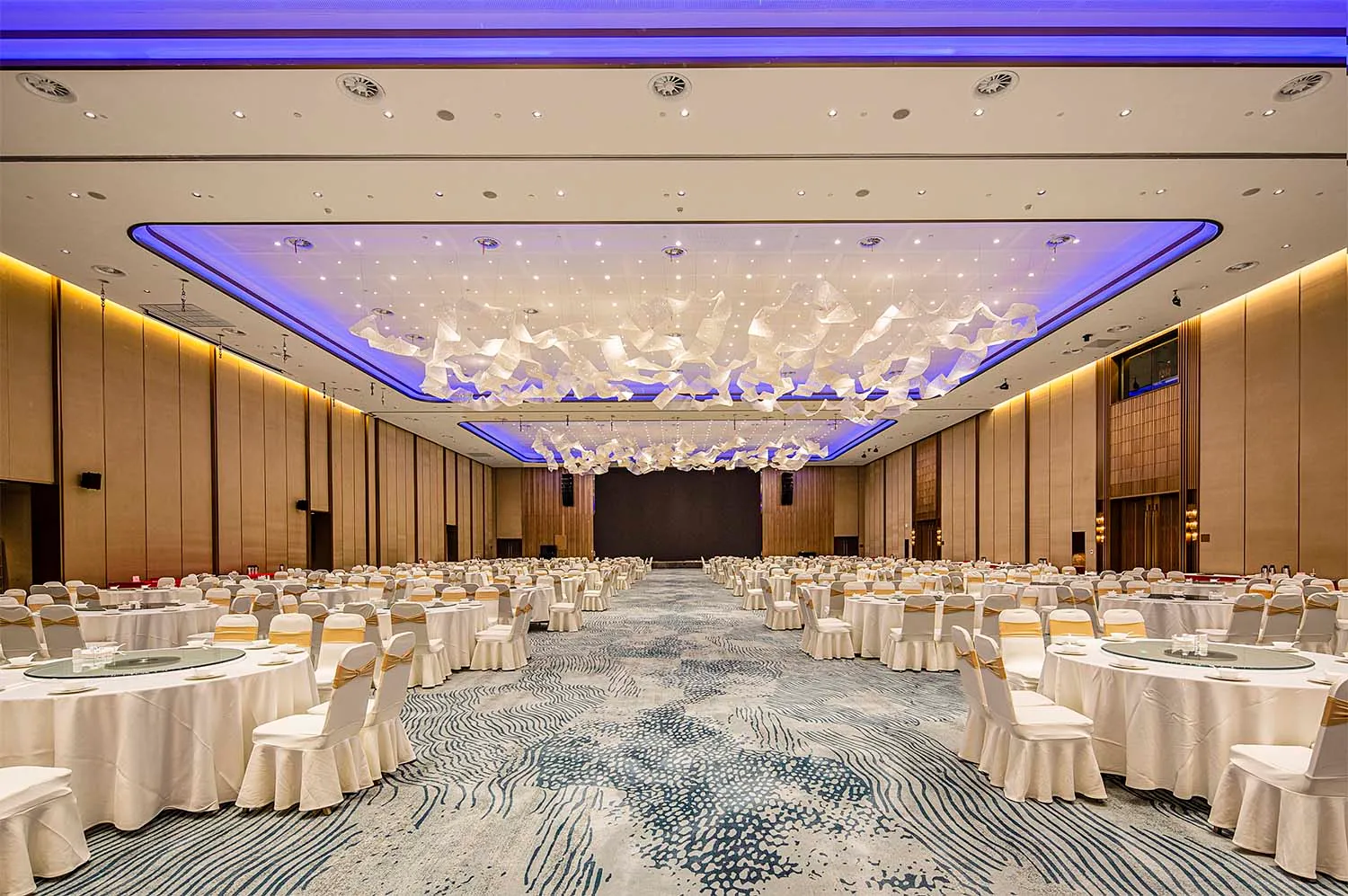 A beautifully decorated banquet hall with white fabric seats, wooden panels, glass chandeliers, and flower decorations.