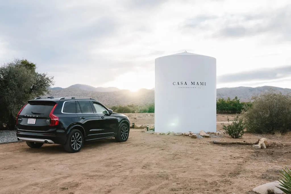 Black SUV parked next to a white cylindrical monument with the words "Casa Mami" on it, against a backdrop of mountains and d