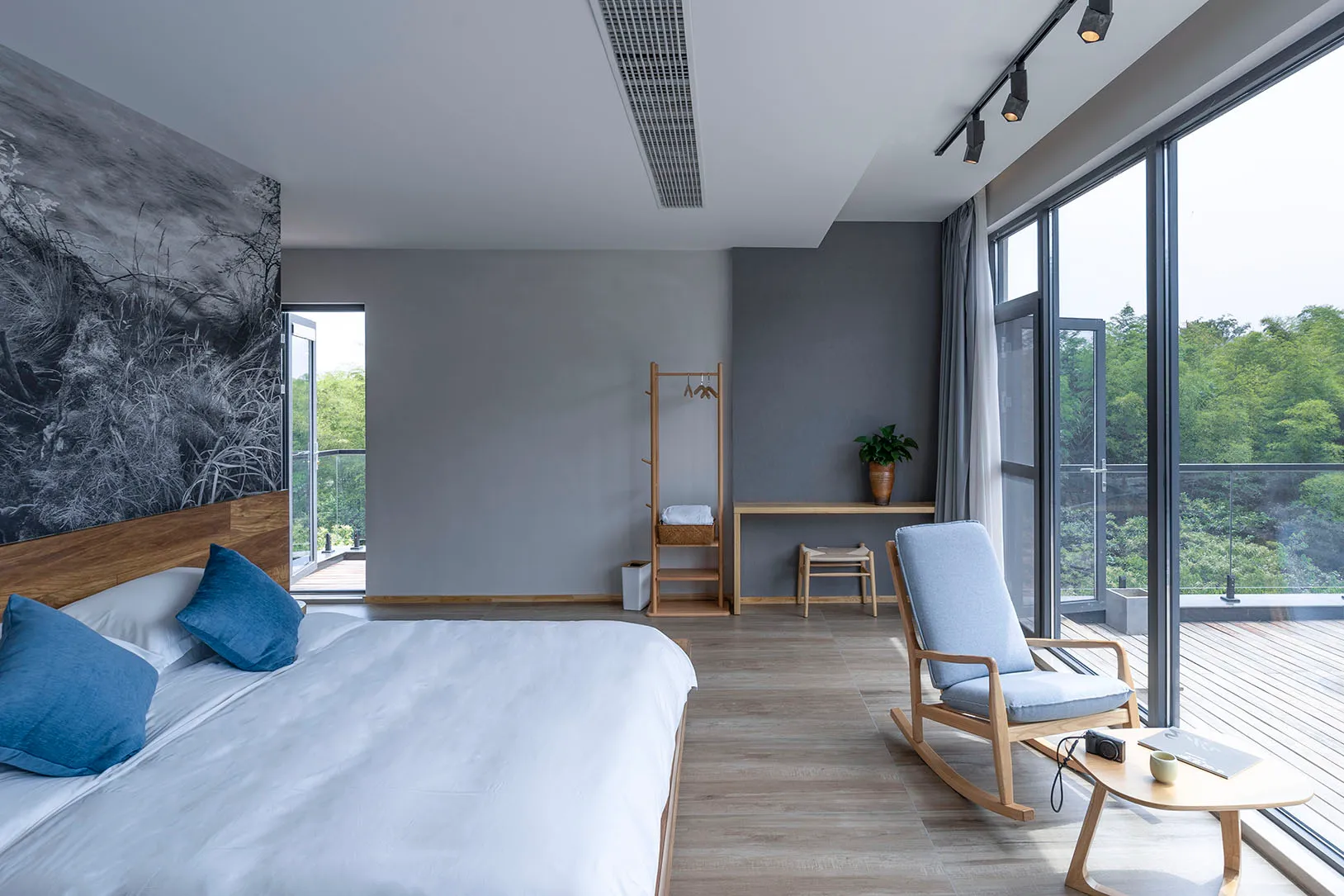 A minimalist bedroom with a large bed, wooden rocking chair, and floor-to-ceiling glass window overlooking greenery.