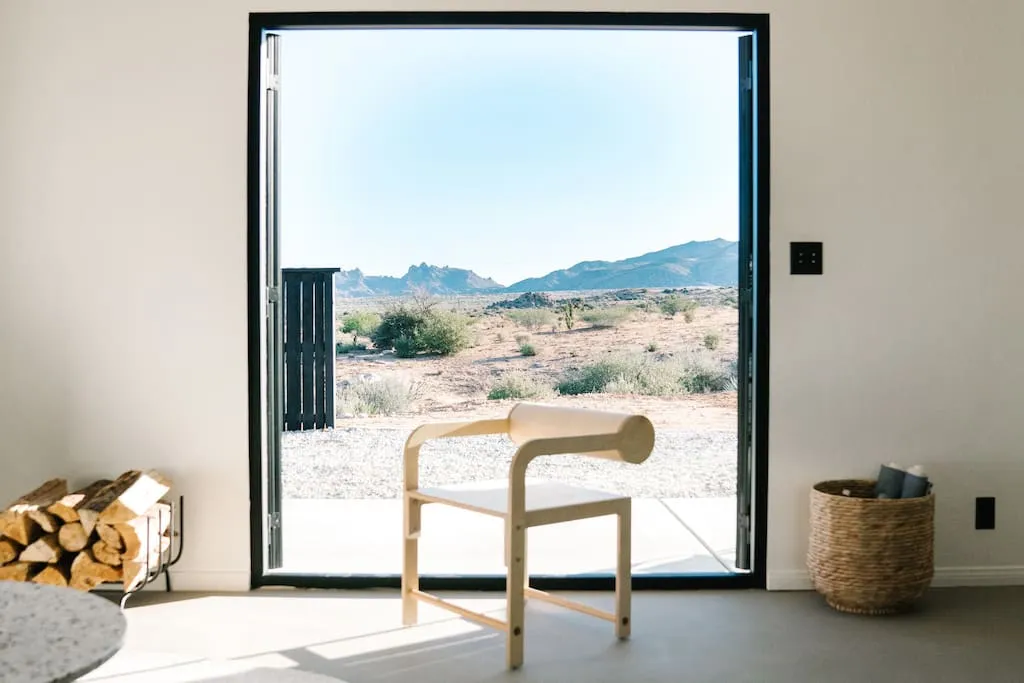 A minimalist living room with a desert view through a large window, featuring a wooden chair, a wicker basket, and a stack of
