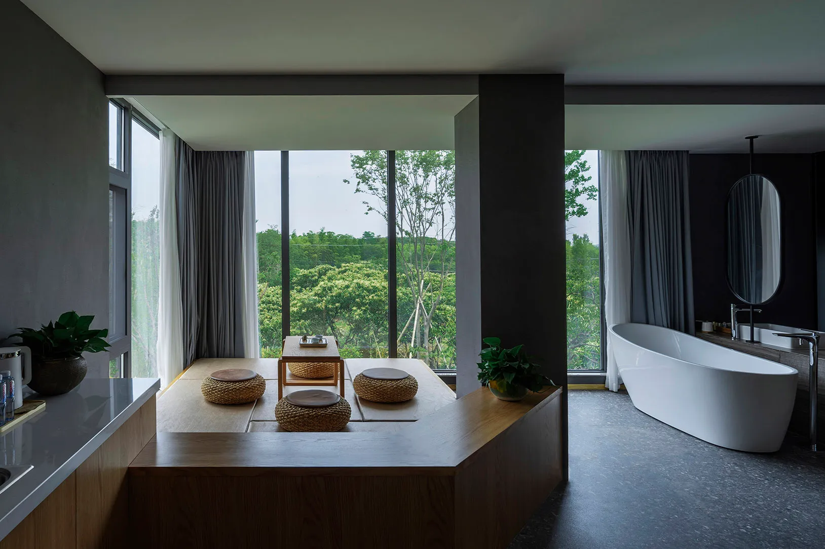 A modern residential bathroom with wood, concrete, and glass elements, featuring a bathtub, seating area, and large windows.