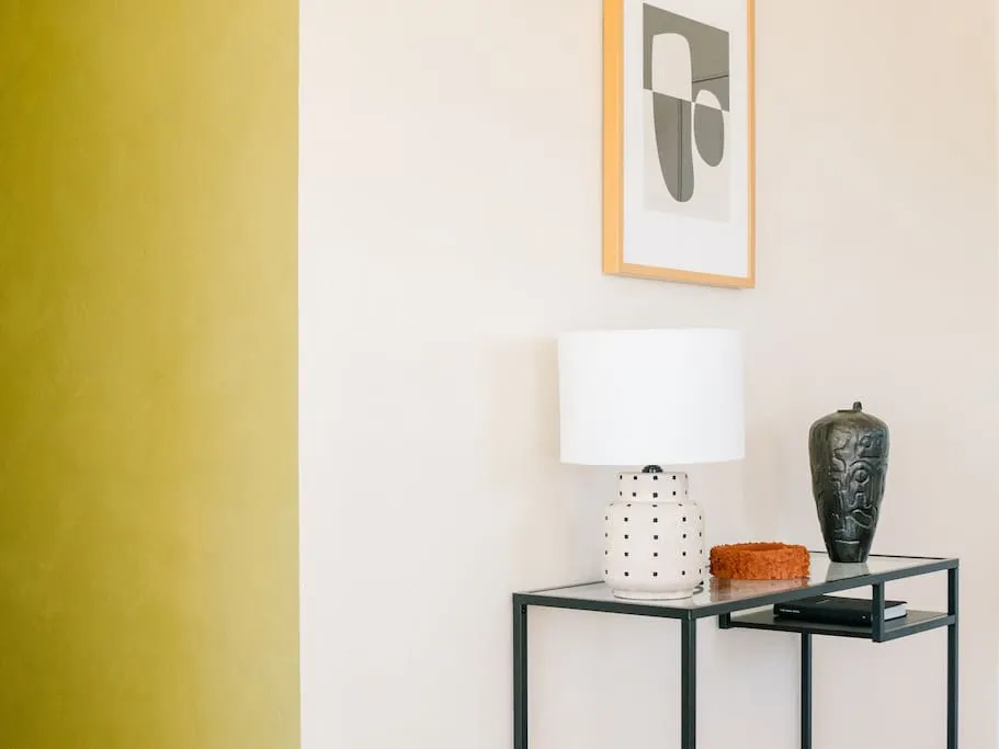 A side table with a modern ceramic lamp and a black and white vase, against a wall with a section painted yellow.