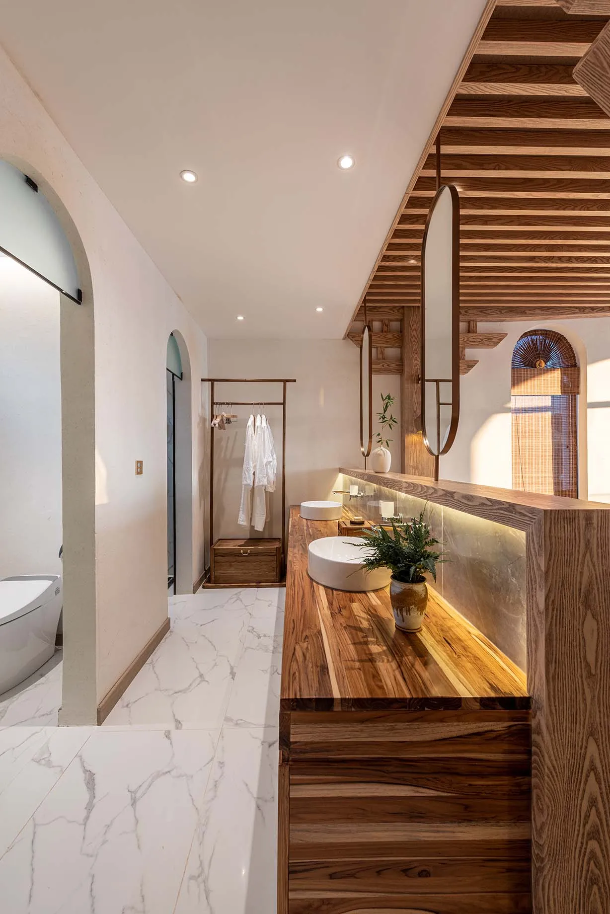 A modern bathroom with white marble flooring, a wooden vanity, and a walk-in closet with hanging robes.