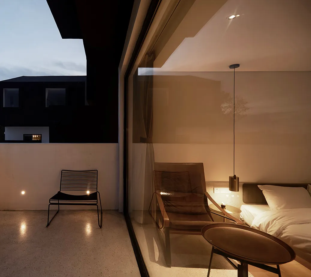 A minimalist bedroom featuring a glass wall, a bed, a chair, and a table with a pendant lamp.