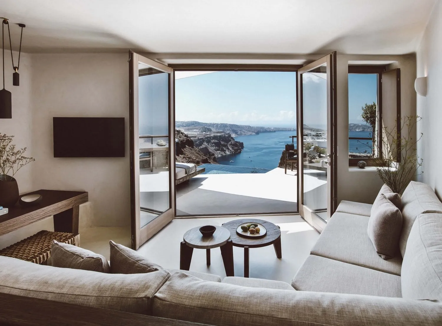 A minimalist living room featuring a sea view, with a large sectional sofa, wooden coffee tables, and a flat-screen TV mounte