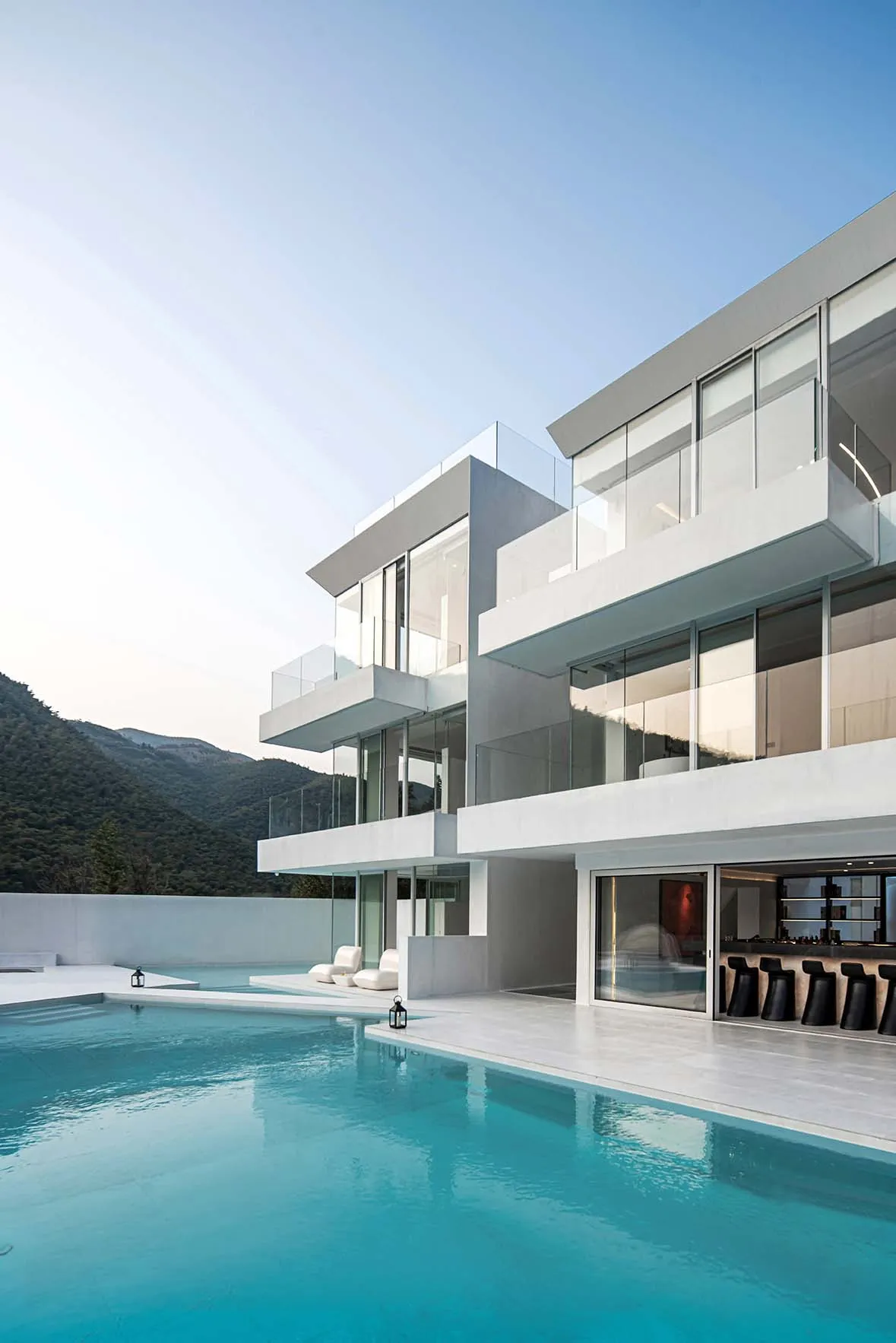 Modern residential house with glass walls, concrete balconies, and a swimming pool overlooking mountains.
