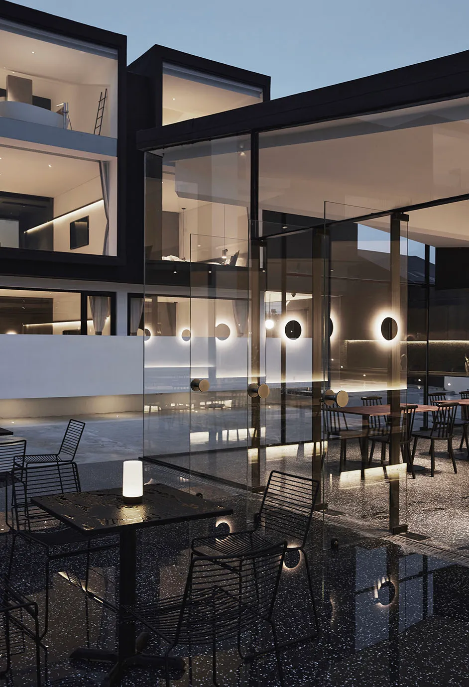 A modern restaurant exterior with glass walls at dusk, illuminated by warm interior lighting.