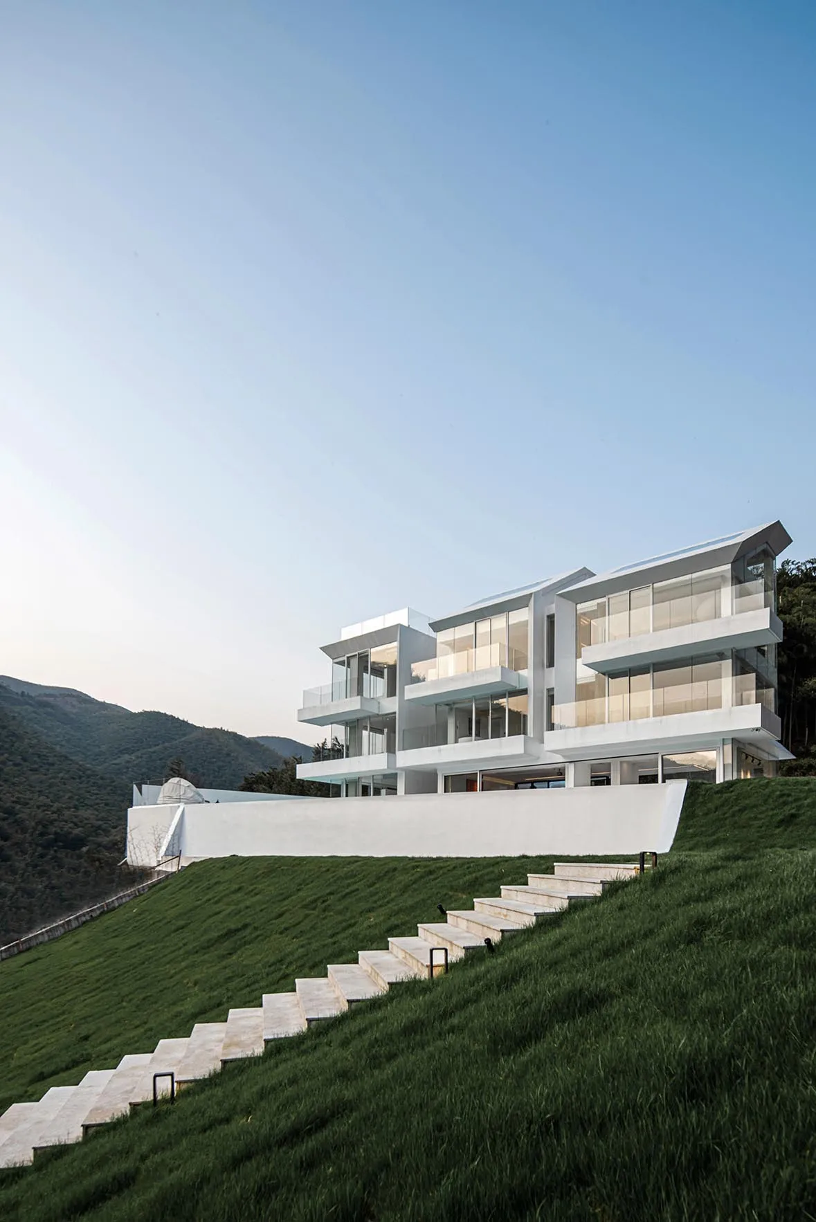A modern residential house built into a lush, green hillside with glass walls and white concrete materials.
