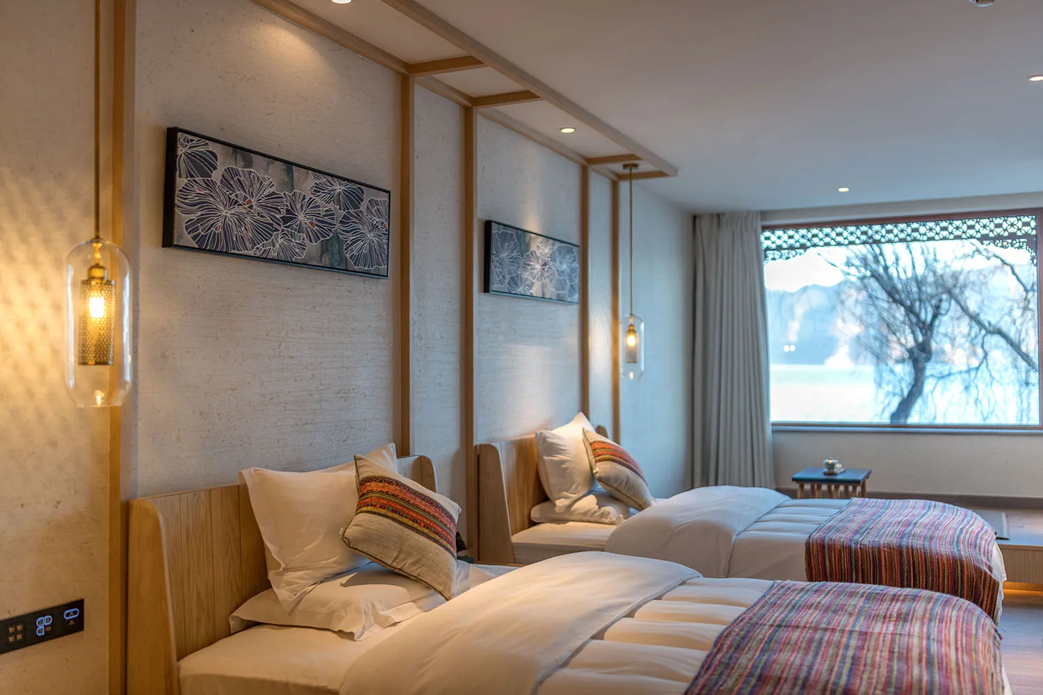 A modern bedroom with textured wall, two twin beds, decorative pillows, and a large window with a view of a tree.