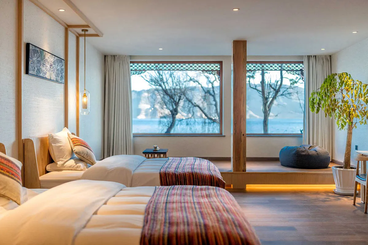 Modern bedroom with two beds, wooden flooring, and large windows showing a scenic view.
