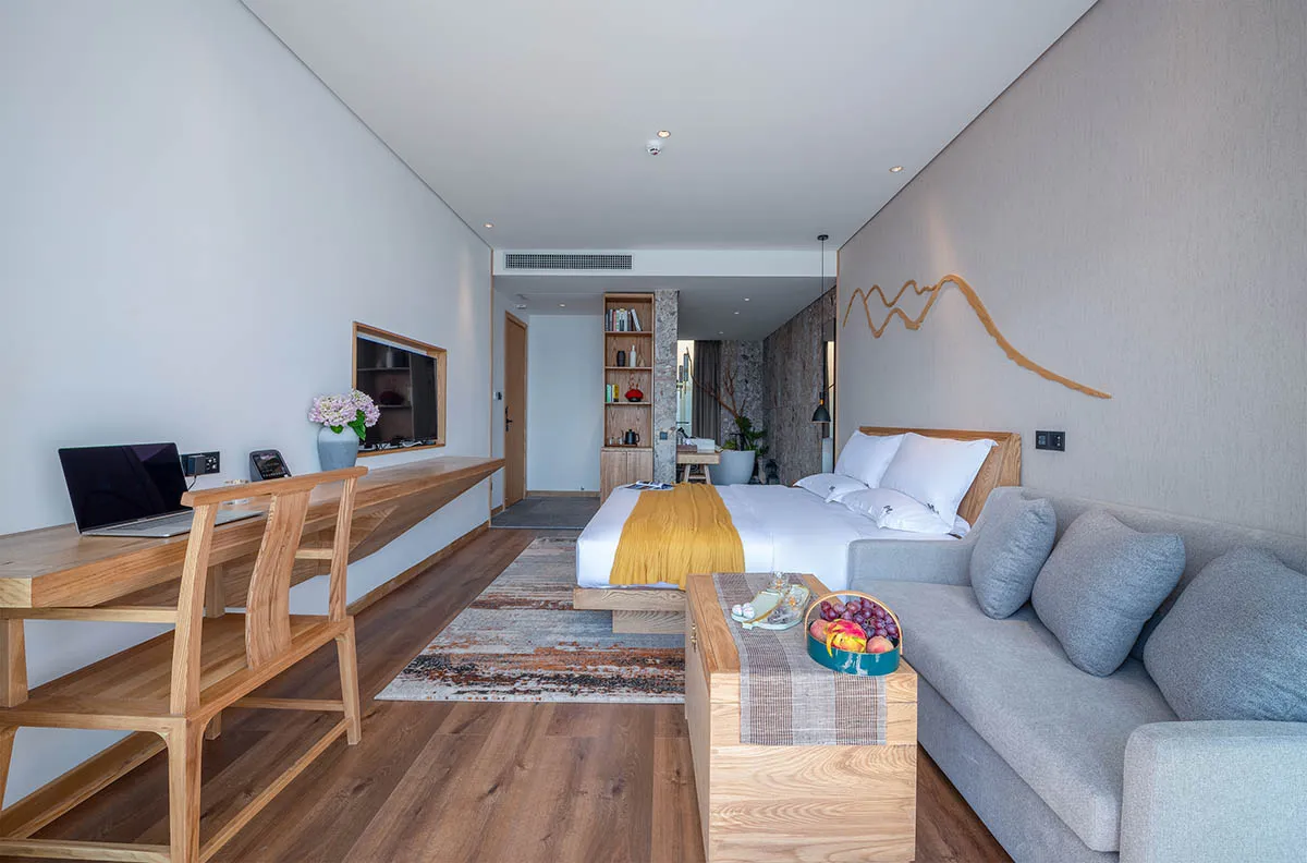 A modern hotel room with minimalist design features, including a wood desk, gray sofa, and a king-sized bed with white beddin