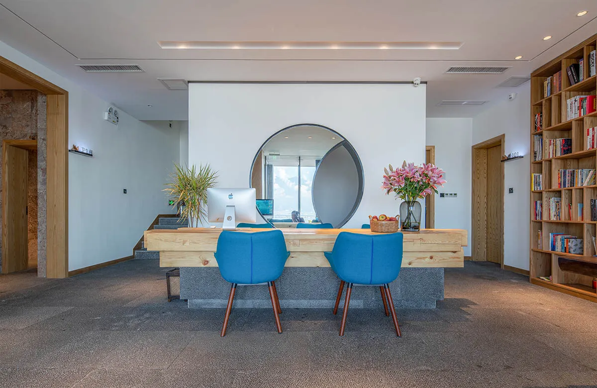 A modern reception desk made of wood, featuring blue chairs and a circular mirror.