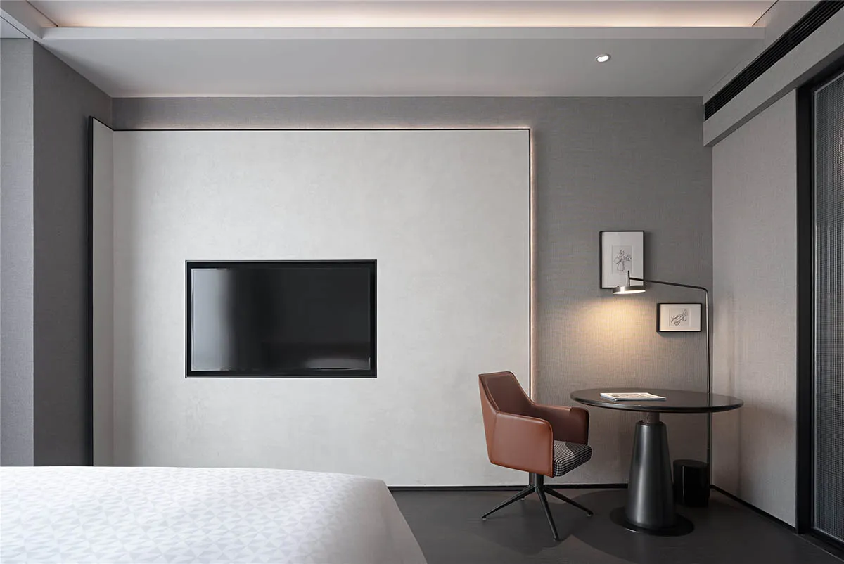 A minimalist bedroom featuring a black leather chair, a flat-screen television mounted on the wall, and a small round table w