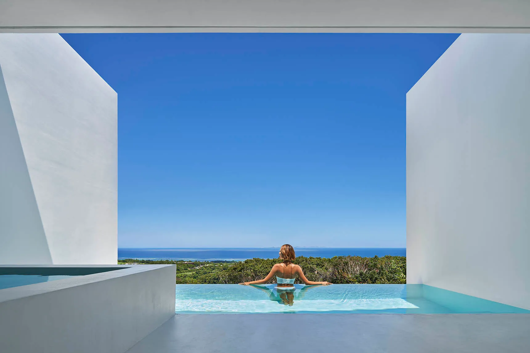A minimalist pool with concrete walls and a clear view of the ocean and trees in the background.