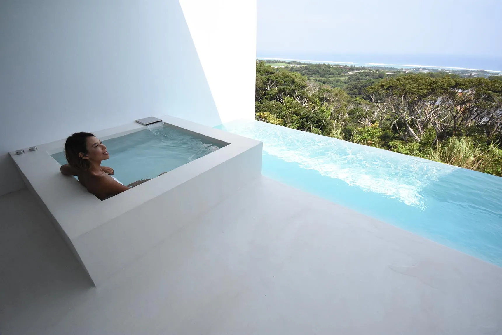 A woman relaxing in a minimalist concrete outdoor spa with scenic views of the forest and ocean.