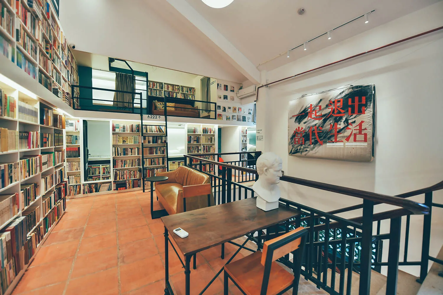 Library interior with concrete walls, metal railings, wooden tables and chairs, and concrete floor covered with books.