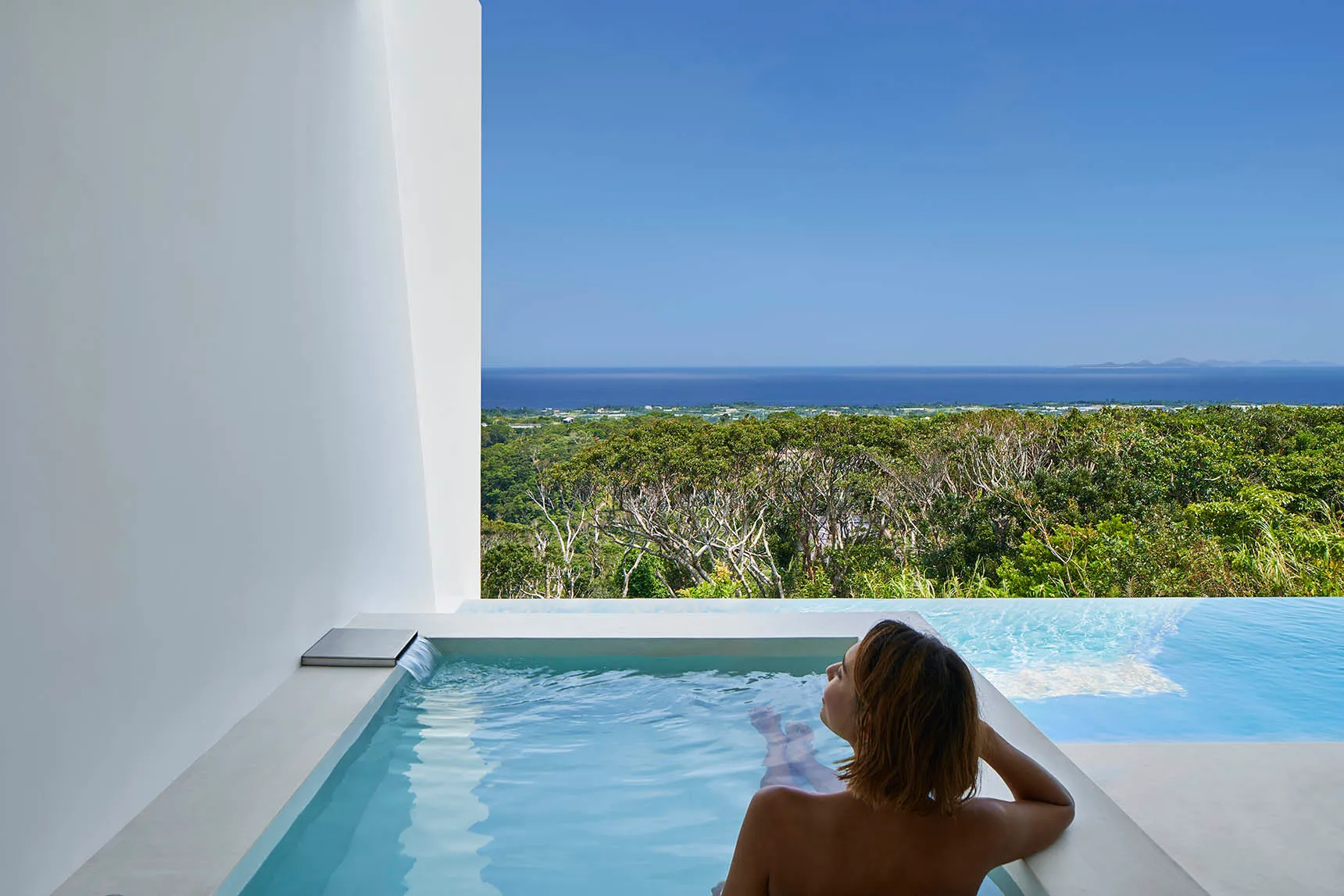 Person relaxing in a modern, minimalist hot tub overlooking lush greenery and the ocean.
