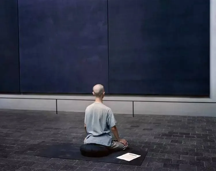 A minimalist installation art piece consisting of a person sitting on a cork mat and looking at a piece of paper in a museum 