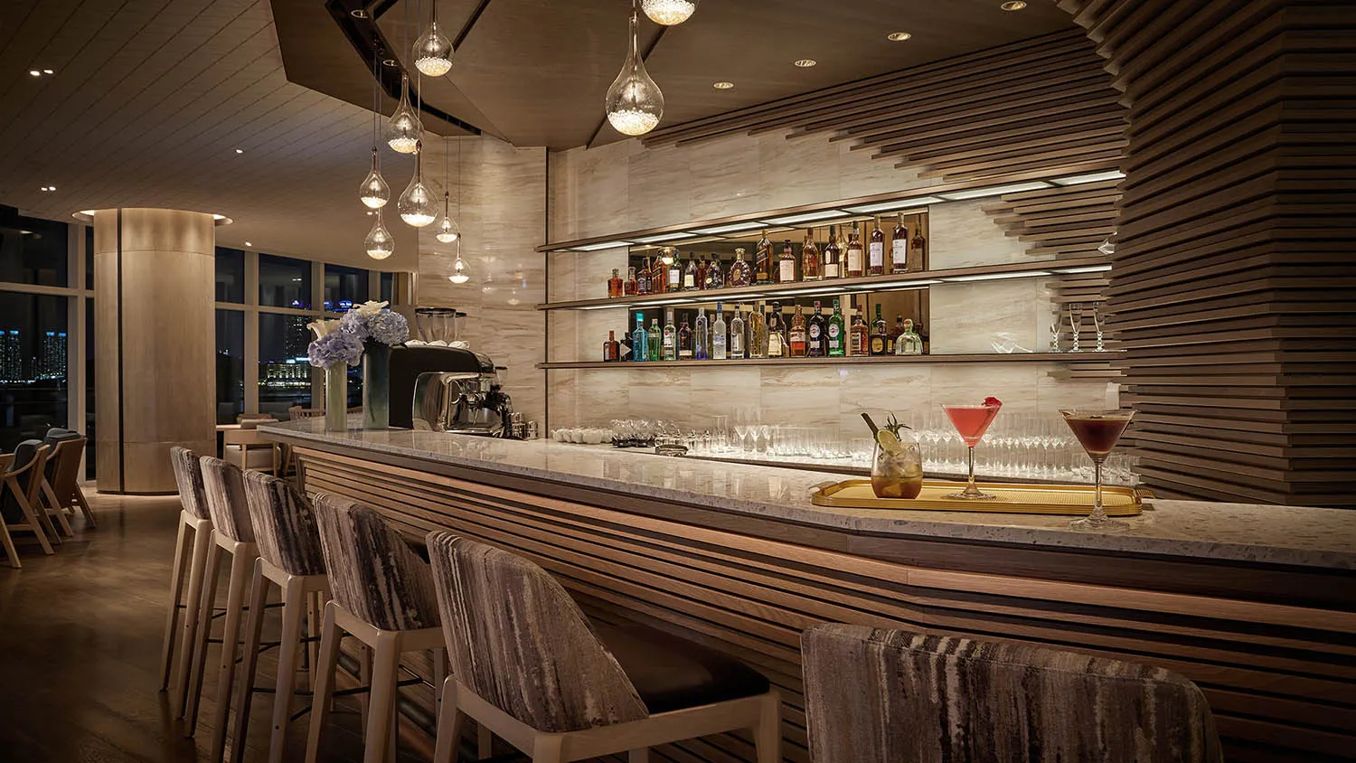 A modern bar with marble and wooden decor, featuring a well-stocked bar counter, chairs, and hanging lights.