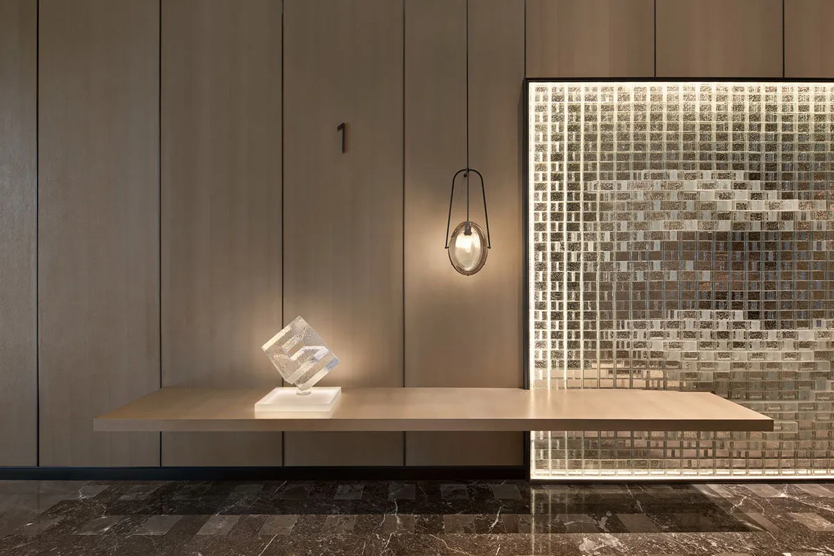 A modern wooden floating desk with a geometric art piece on top, illuminated by a pendant light and a mosaic wall backdrop.