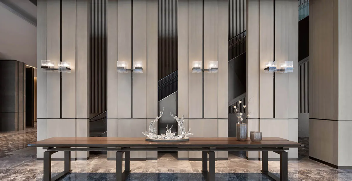 A modern lobby features a long wooden table with metal legs, adorned with a glass water feature, set against a backdrop of ma