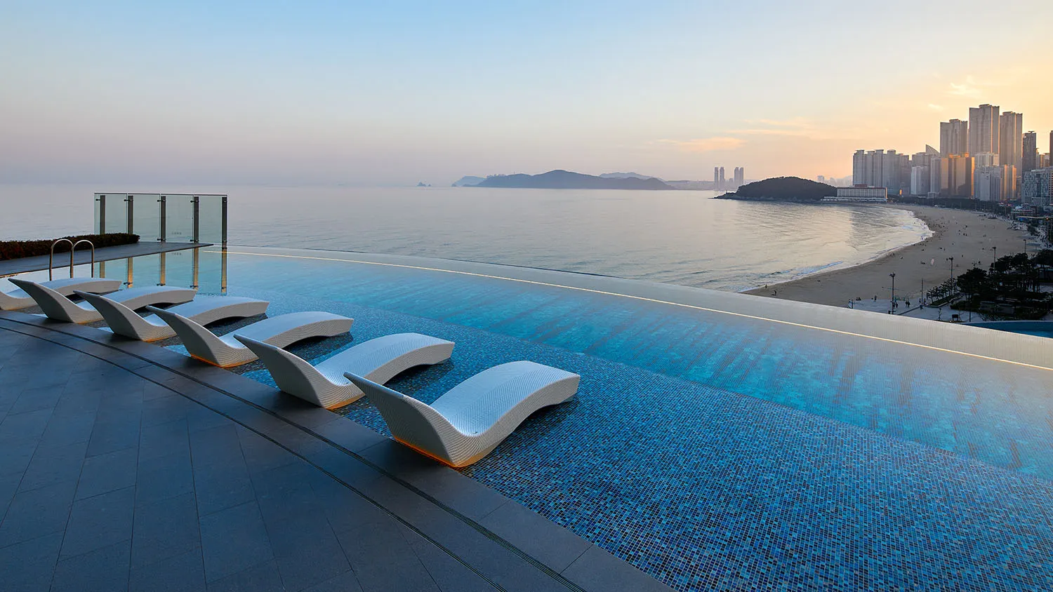 An infinity pool with glass walls and modern seating overlooking a city and the sea.