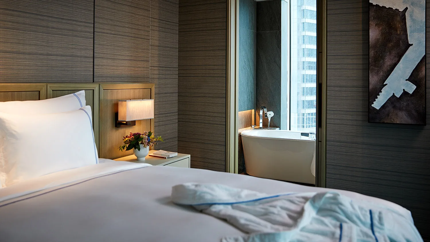 Modern bedroom with wooden headboard and bedside table, white bedding, and a bathtub with a city view.