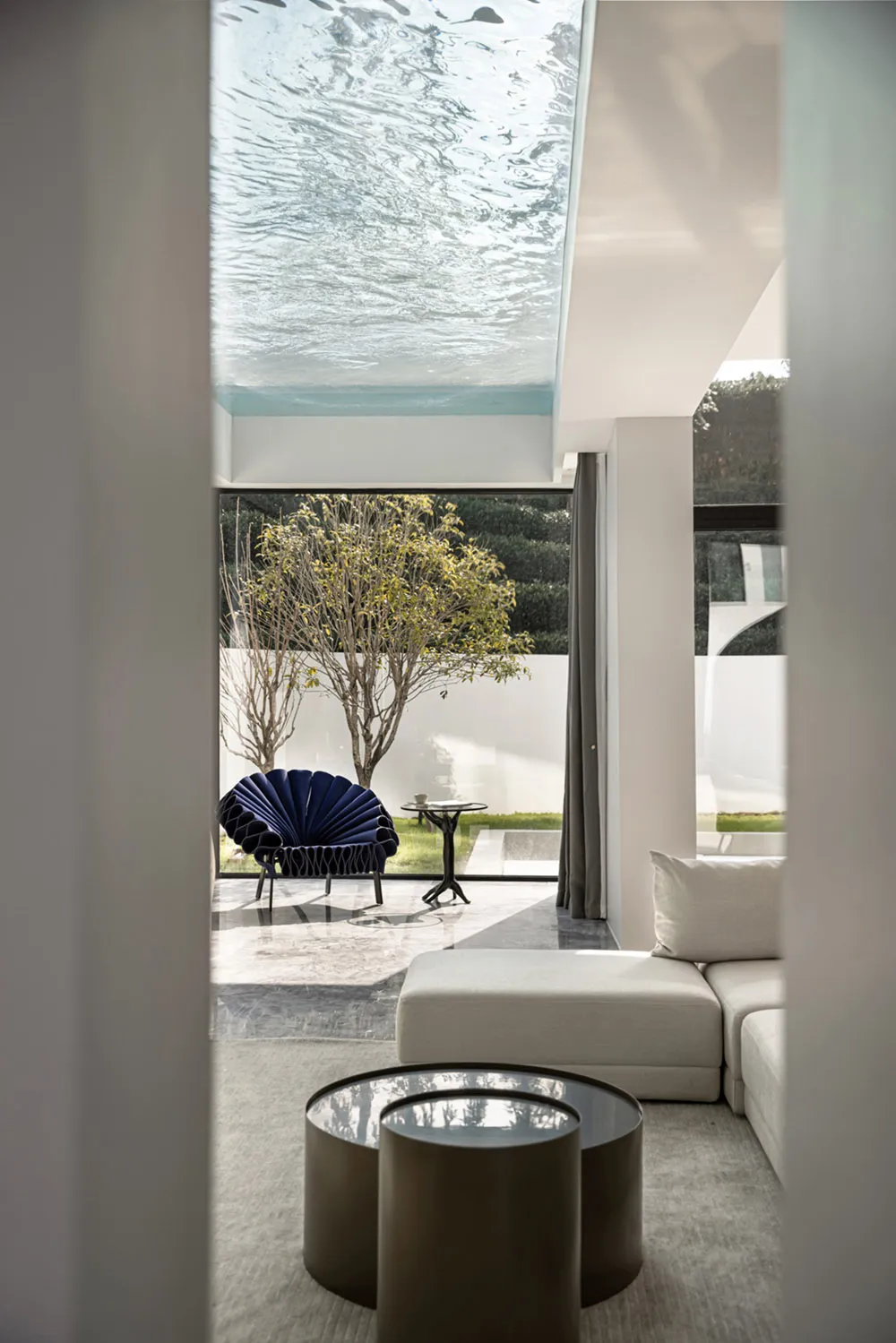 A modern living room with a glass ceiling, featuring a white sectional sofa, blue accent chair, and a small round table.